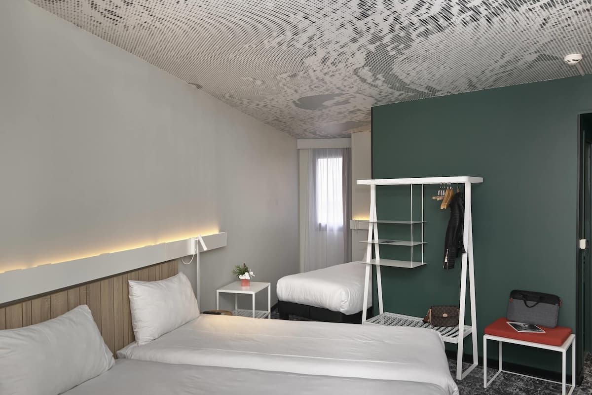 A hotel room with two beds, a white bedside table with a lamp, a small table with a plant, and a coat rack with a jacket and bag.