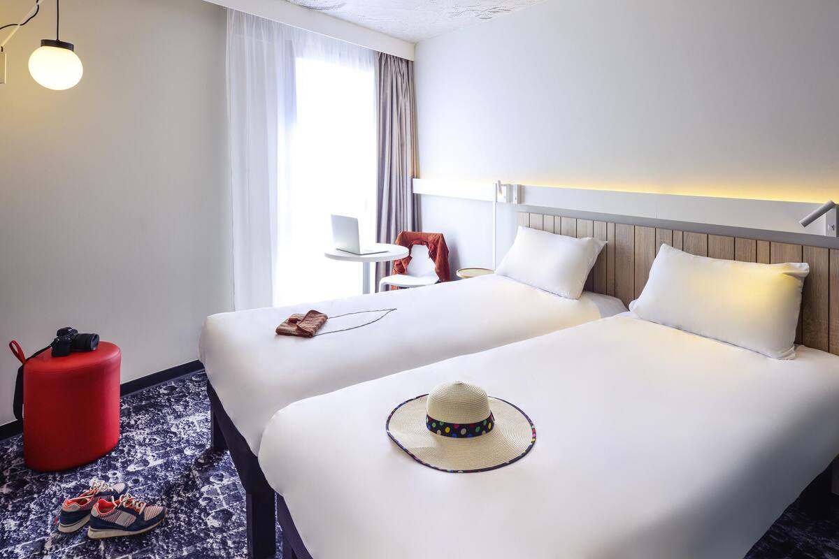 A hotel room with two beds, a red ottoman, a laptop on a small table, and a hat on one of the beds.