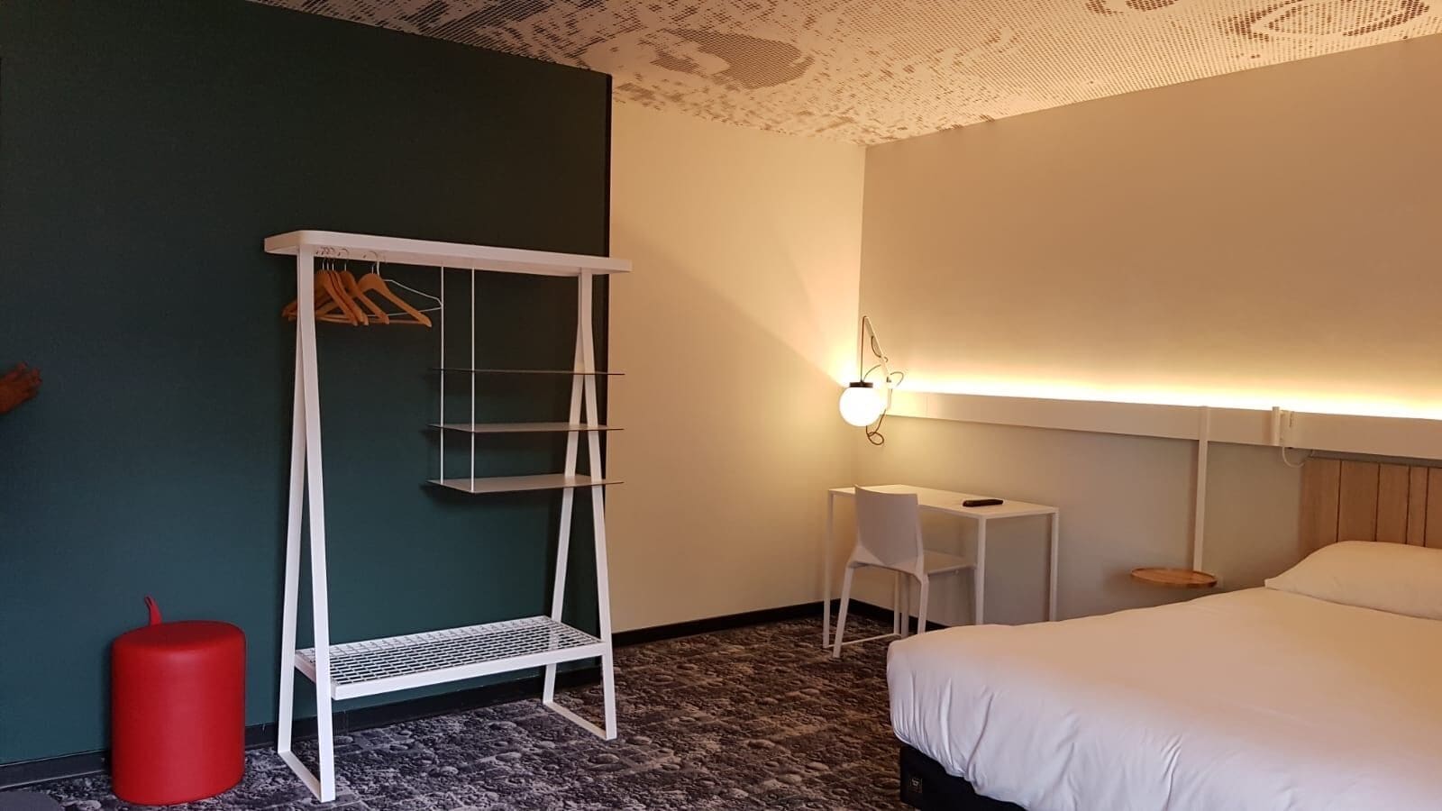Habitación de hotel con cama, escritorio, silla, armario y un cubo de basura rojo.