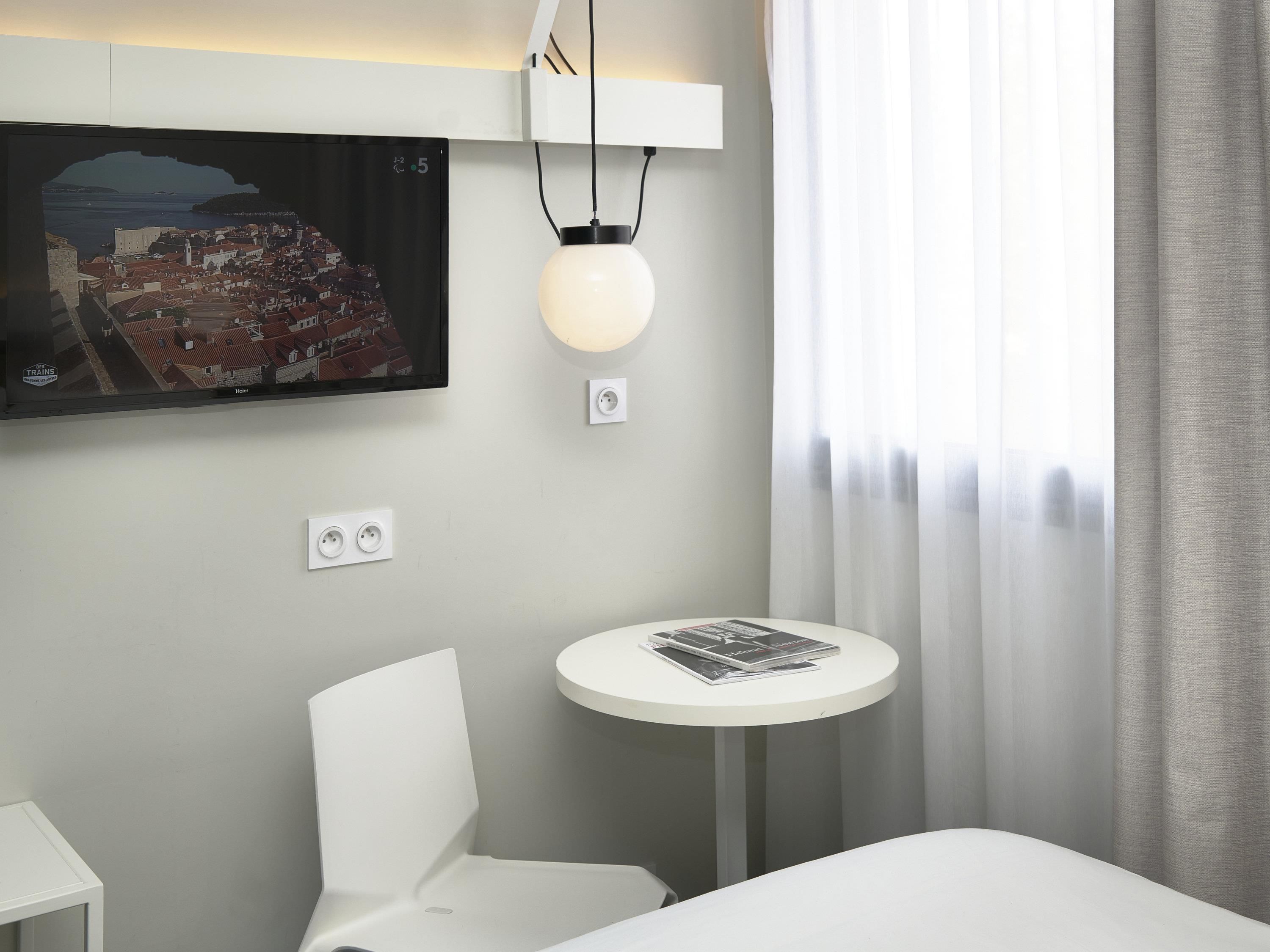 A hotel room with a bed, a white chair, a small round table with magazines, a wall-mounted TV displaying a cityscape, and a window with sheer curtains.