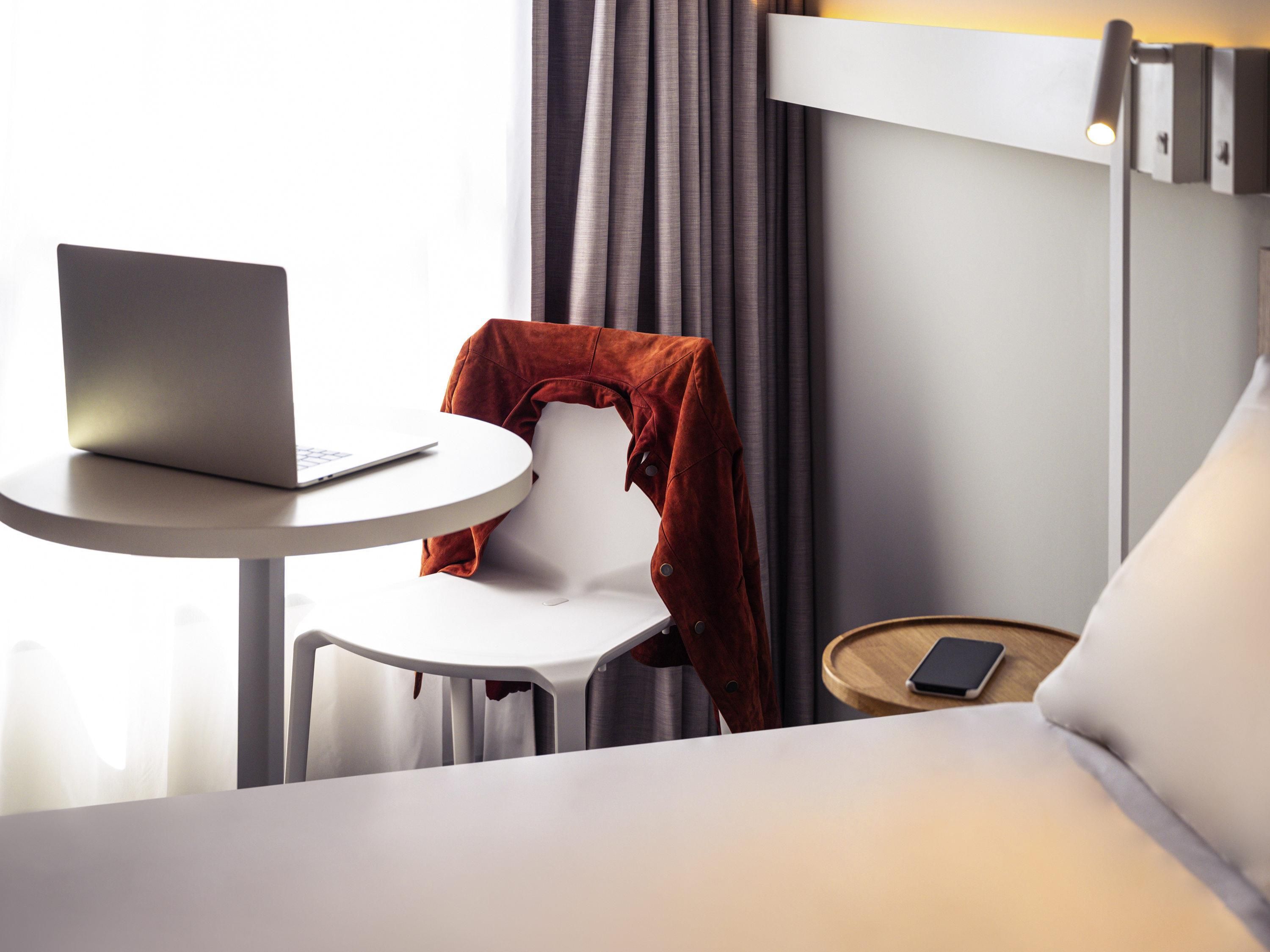 Ein ordentlich eingerichtetes Hotelzimmer mit einem Bett, einem kleinen runden Tisch mit einem Laptop, einem Sessel mit einer Jacke, einem Nachttisch mit einem Telefon und einem Fenster mit Vorhängen.