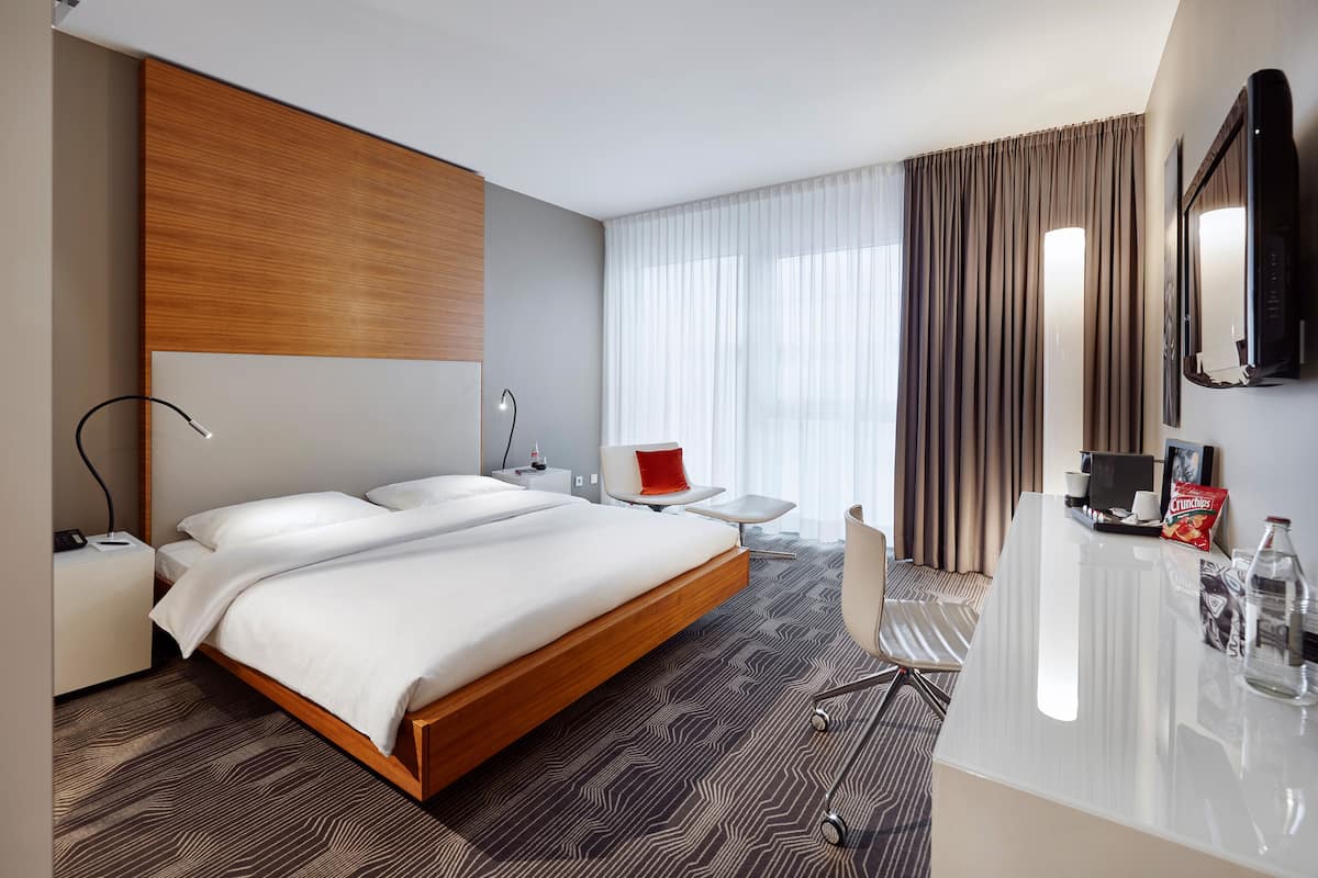 Ein modernes Hotelzimmer mit einem großen Bett, einem Schreibtisch, einem Sessel, einem Fernseher und einer Minibar.