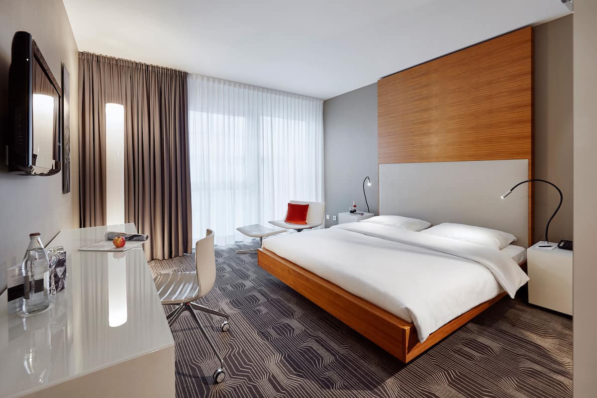 Ein modernes Hotelzimmer mit einem großen Bett, einem Schreibtisch mit Spiegel, einem Sessel, einem Nachttisch mit Lampe und einem großen Fenster mit Vorhängen.
