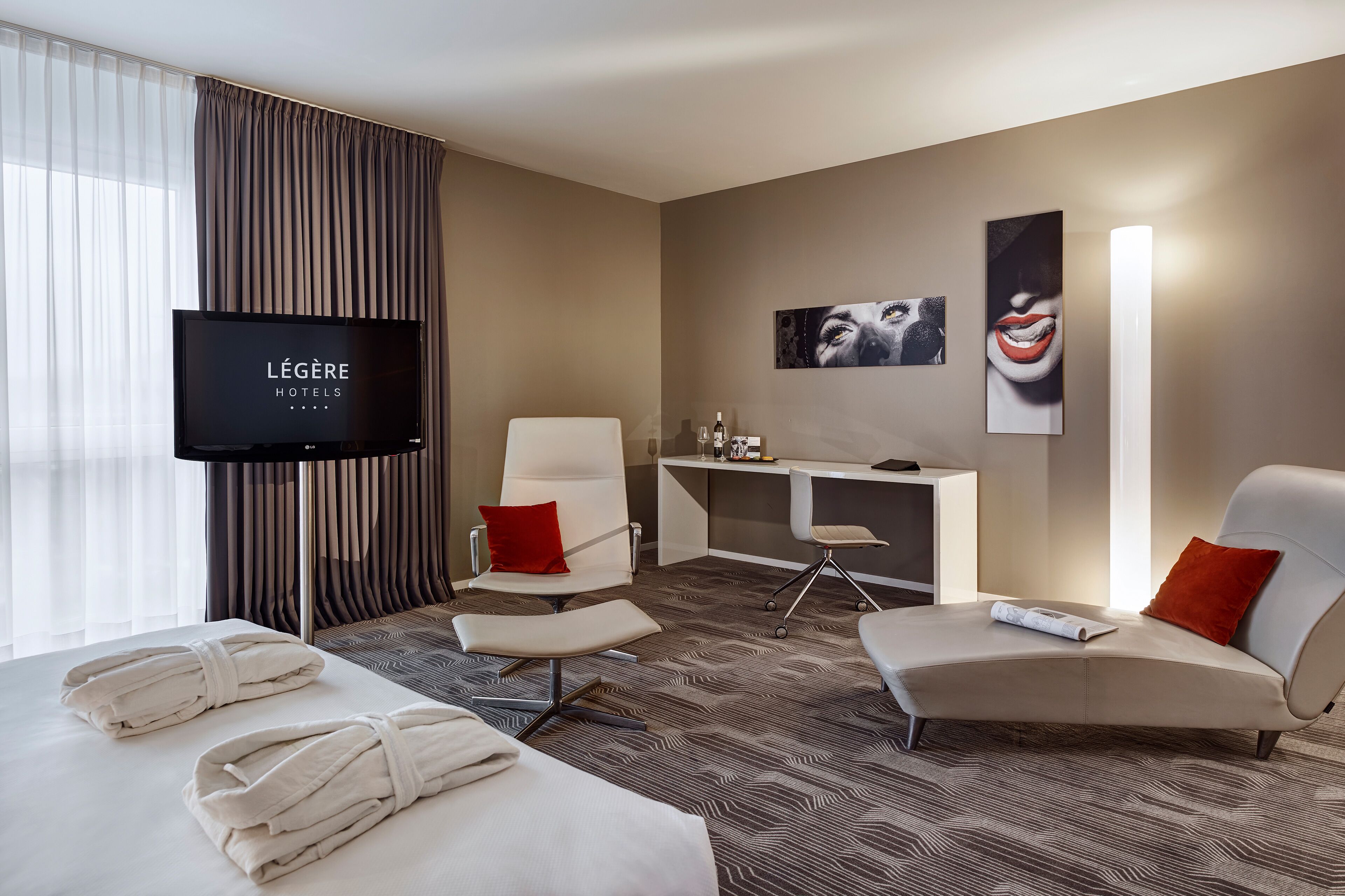 A hotel room with a bed, two armchairs, a desk, a TV, and artwork on the wall.