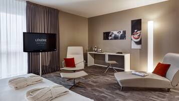 A hotel room with a bed, two armchairs, a desk, a TV, and artwork on the wall.