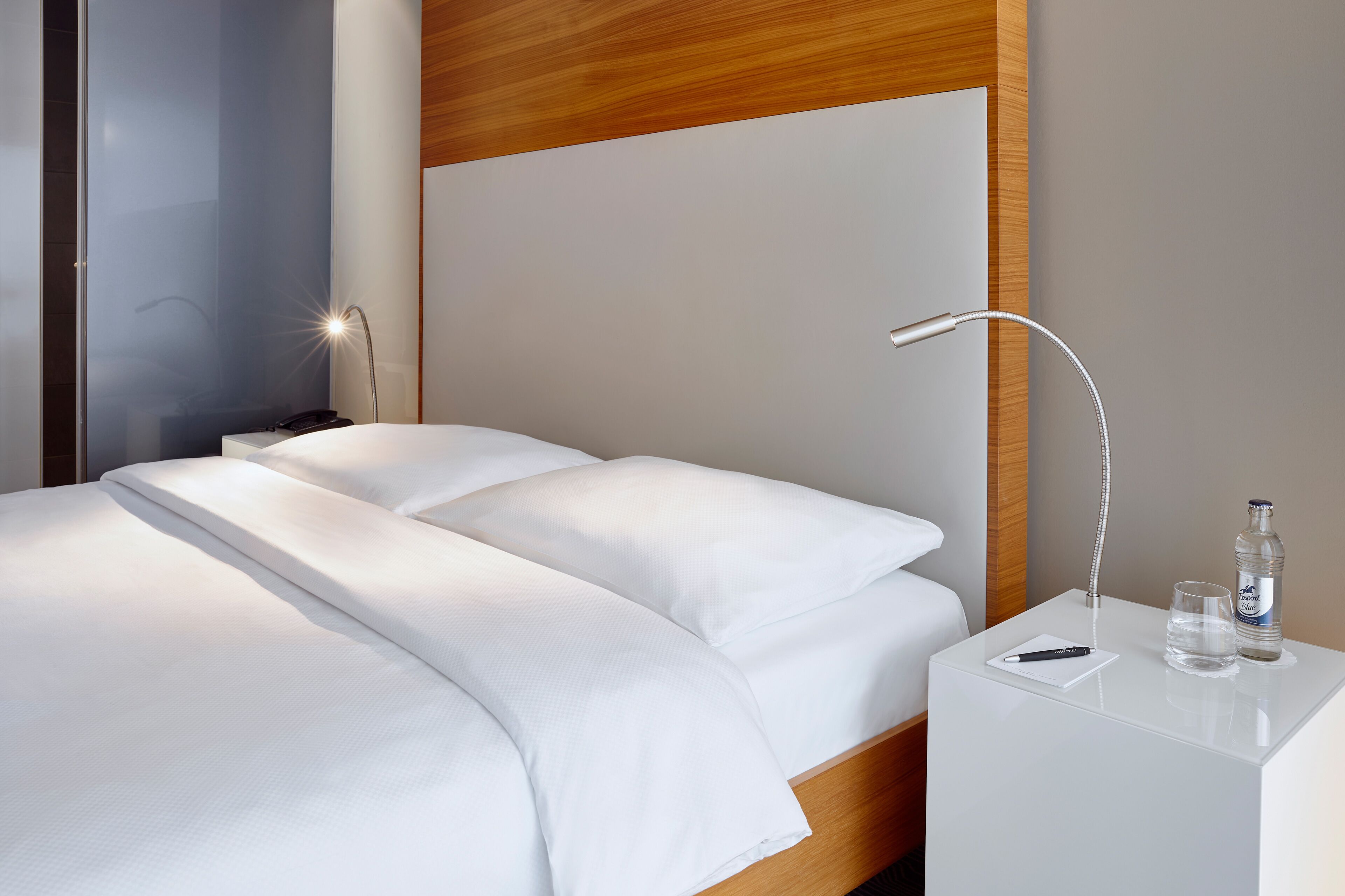 A modern hotel room with a neatly made bed, a bedside table with a lamp, a bottle of water, and a glass.