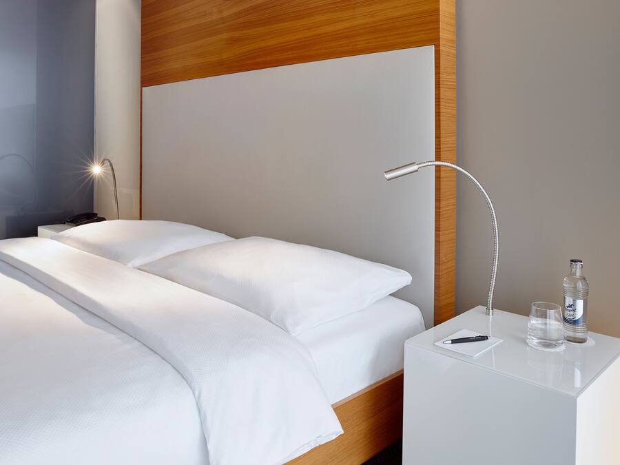 Una habitación de hotel moderna con una cama bien hecha, una mesita de noche con una lámpara, una botella de agua y un vaso.