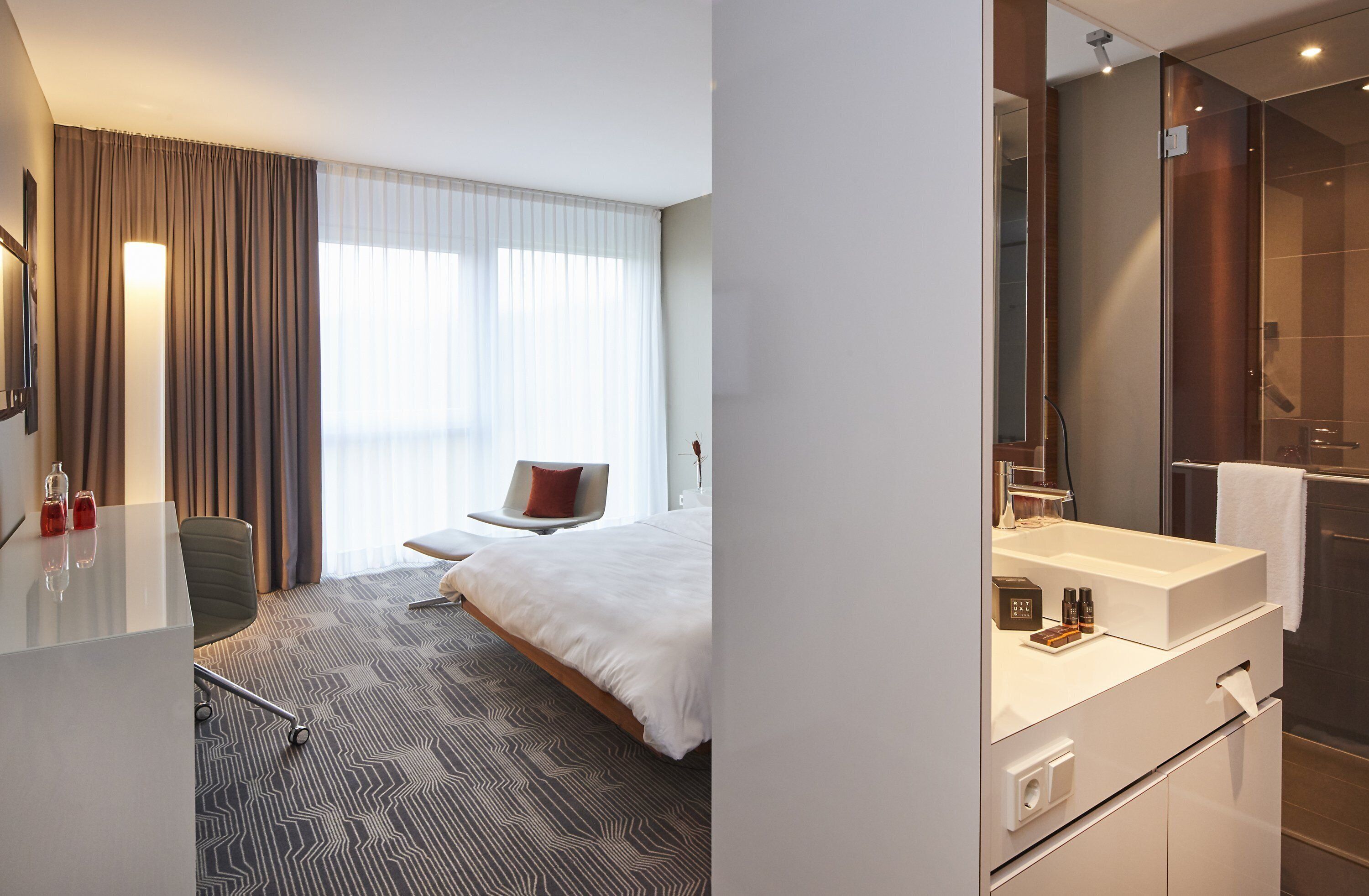 A modern hotel room with a large bed, a desk, a chair, a bathroom with a large mirror, and a shower.
