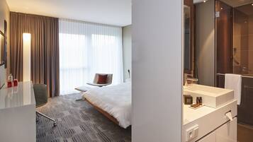 A modern hotel room with a large bed, a desk, a chair, a bathroom with a large mirror, and a shower.
