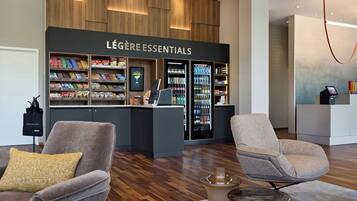 A hotel lobby with a Légère Hotel Group store, featuring a variety of snacks and beverages, two grey armchairs, and a modern light fixture.