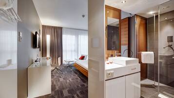 Una habitación de hotel moderna con cabina de ducha de vidrio, un espejo grande, una mesita de noche y una cama con almohada roja.