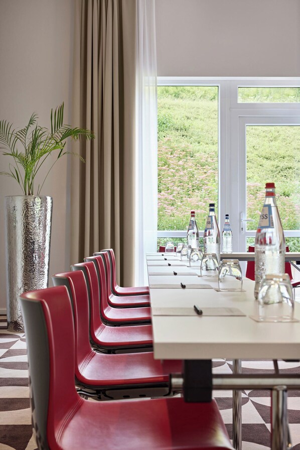A conference room with a long table, red chairs, a potted plant, and a view of greenery through the window. - LEGERE HOTEL Luxembourg (Schuttrange)