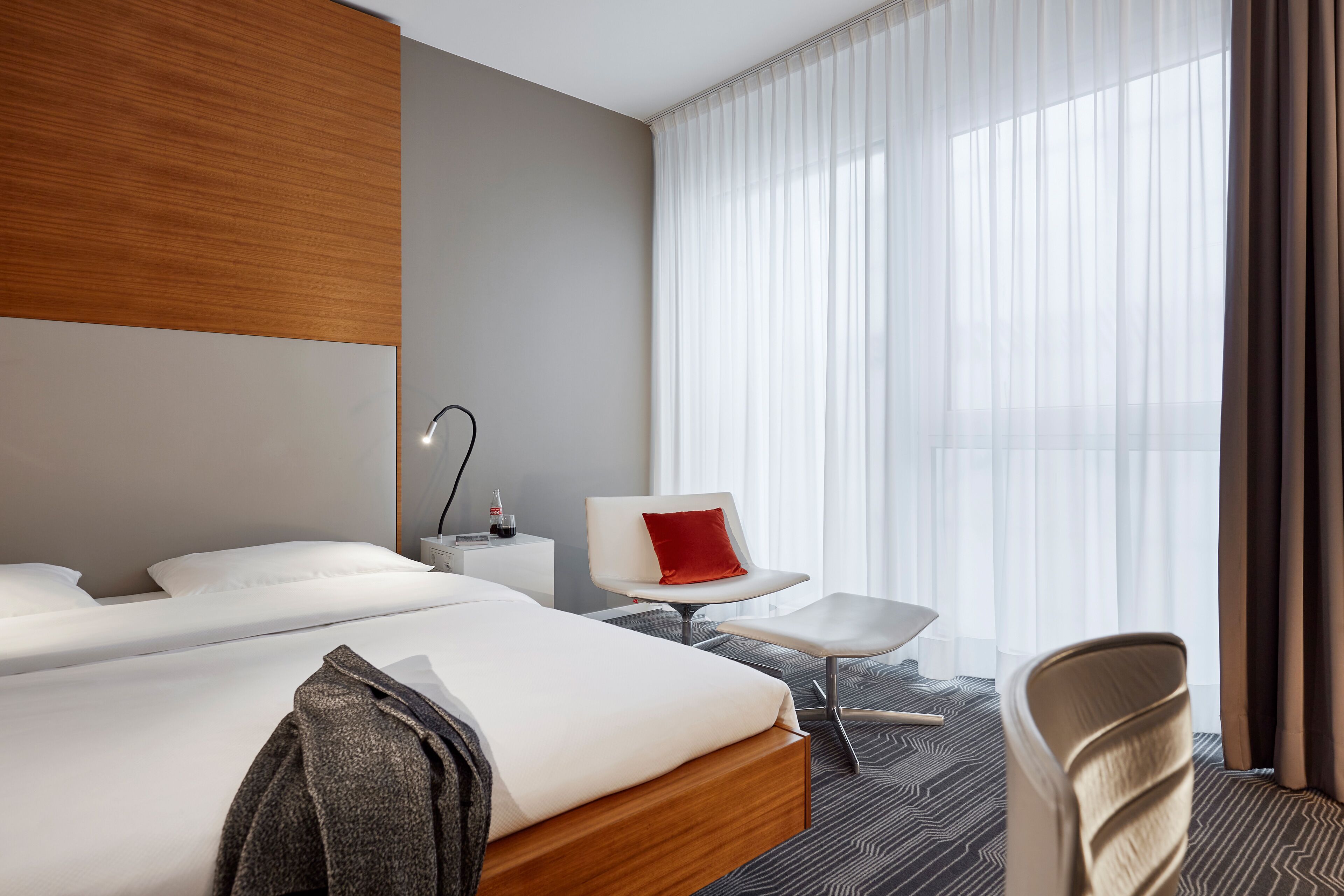Quarto de hotel moderno com duas camas, uma escrivaninha, uma cadeira e uma janela grande com cortinas.