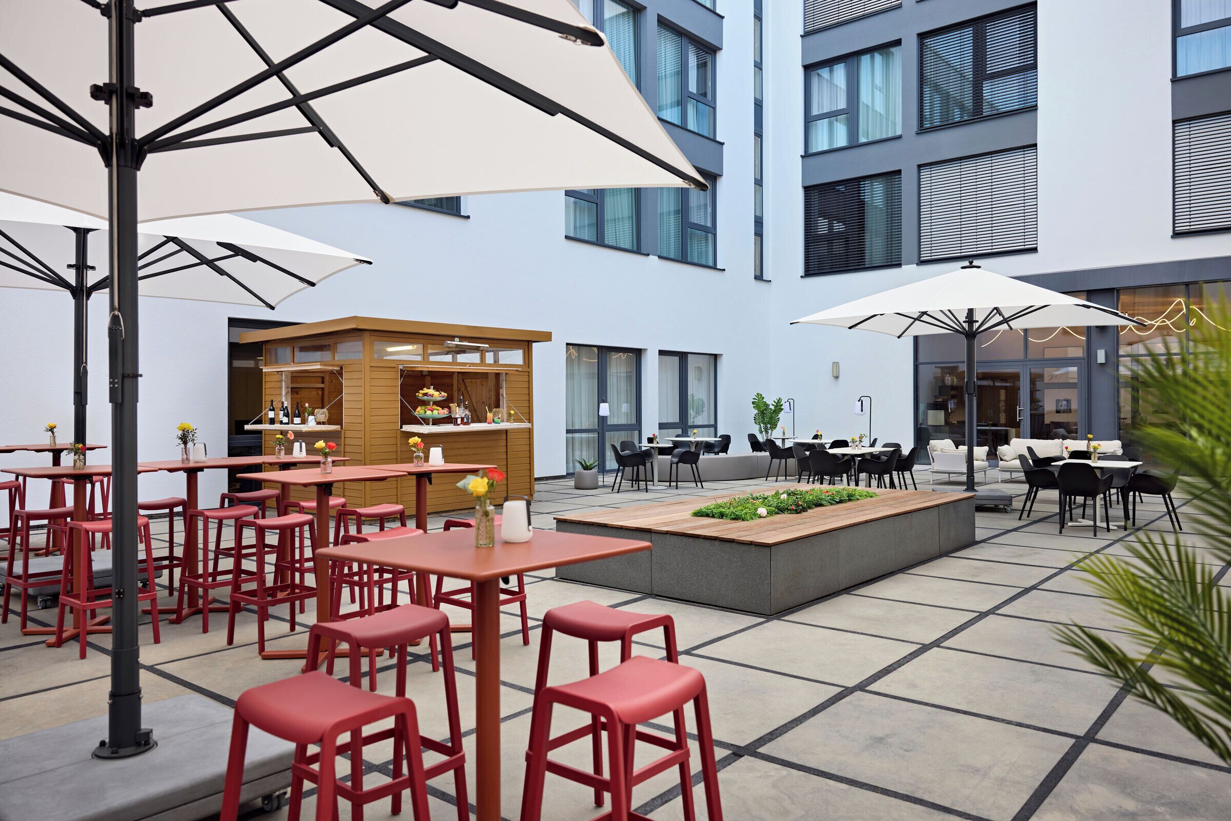 A modern outdoor seating area with red chairs and tables, umbrellas, and a central planter.