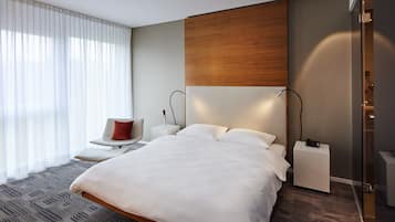 Ein modernes Hotelzimmer mit einem großen Bett, Nachttischen, einem Sessel und Blick auf die Stadt durch große Fenster.