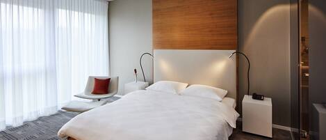A modern hotel room with a large bed, bedside tables, a chair, and a view of the city through large windows.