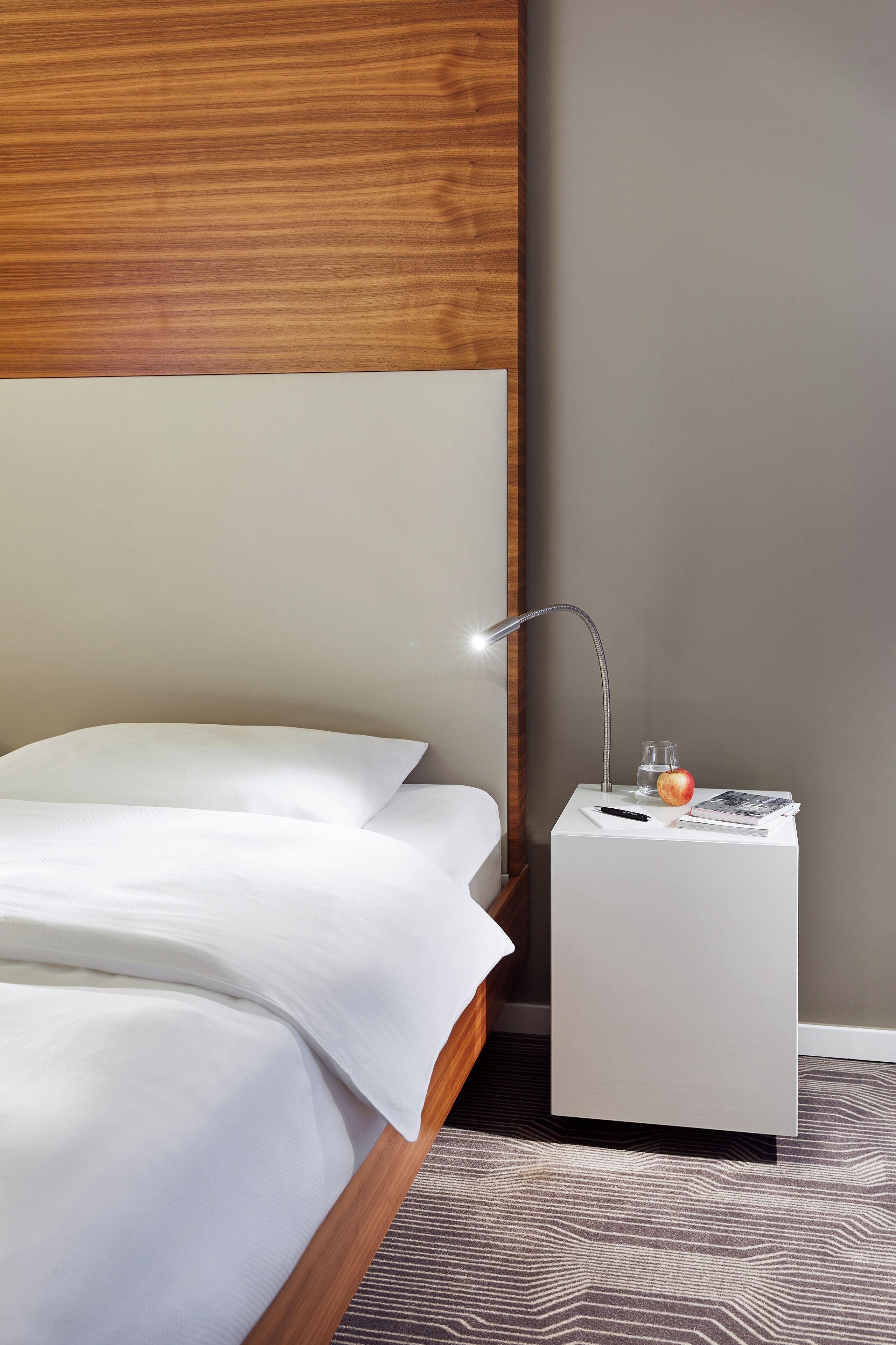 A neatly made bed with white linens, a bedside lamp, a small white nightstand with a glass of water, an apple, and a magazine.