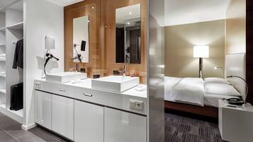 Un baño de hotel moderno con lavamanos dobles, un espejo grande y una zona de dormitorio separada con cama y mesita de noche.