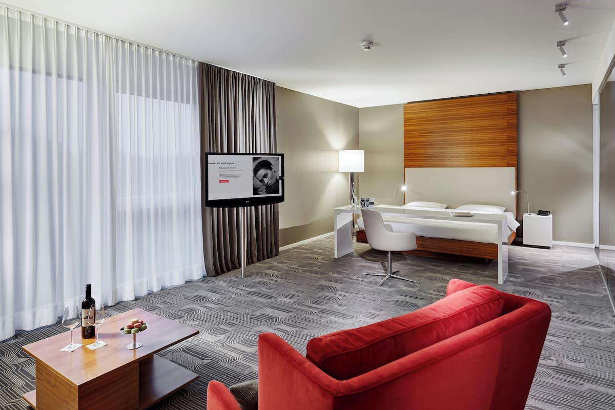 Ein modernes Hotelzimmer mit einem Bett, einem Schreibtisch, einem Sessel, einem roten Sofa, einem Fernseher und einer Weinflasche auf einem Tisch.