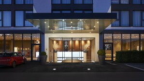 Entrada de un hotel moderno con fachada de vidrio, un coche estacionado afuera y un interior bien iluminado visible a través de las puertas.