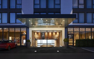 A modern hotel entrance with a glass facade, a car parked outside, and a well-lit interior visible through the doors.