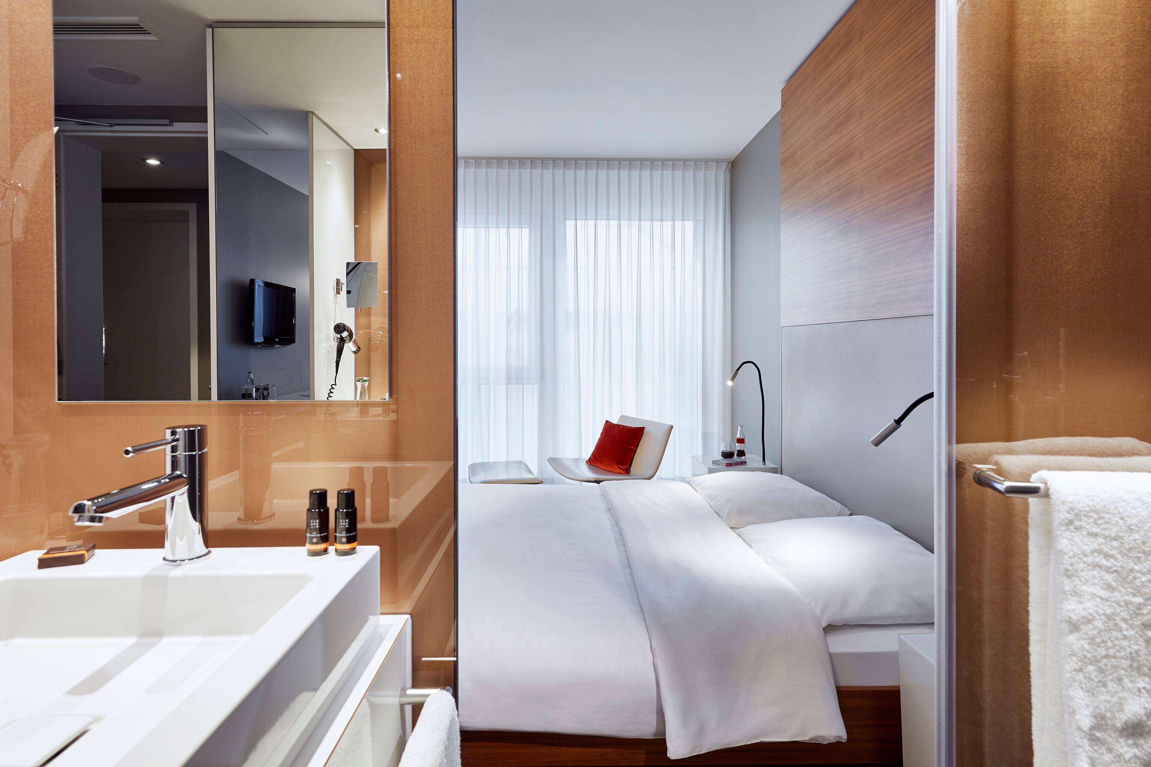 Une chambre d’hôtel moderne avec un grand miroir, un lavabo blanc, un lit avec du linge de lit blanc, un oreiller rouge et un mur recouvert de panneaux en bois.