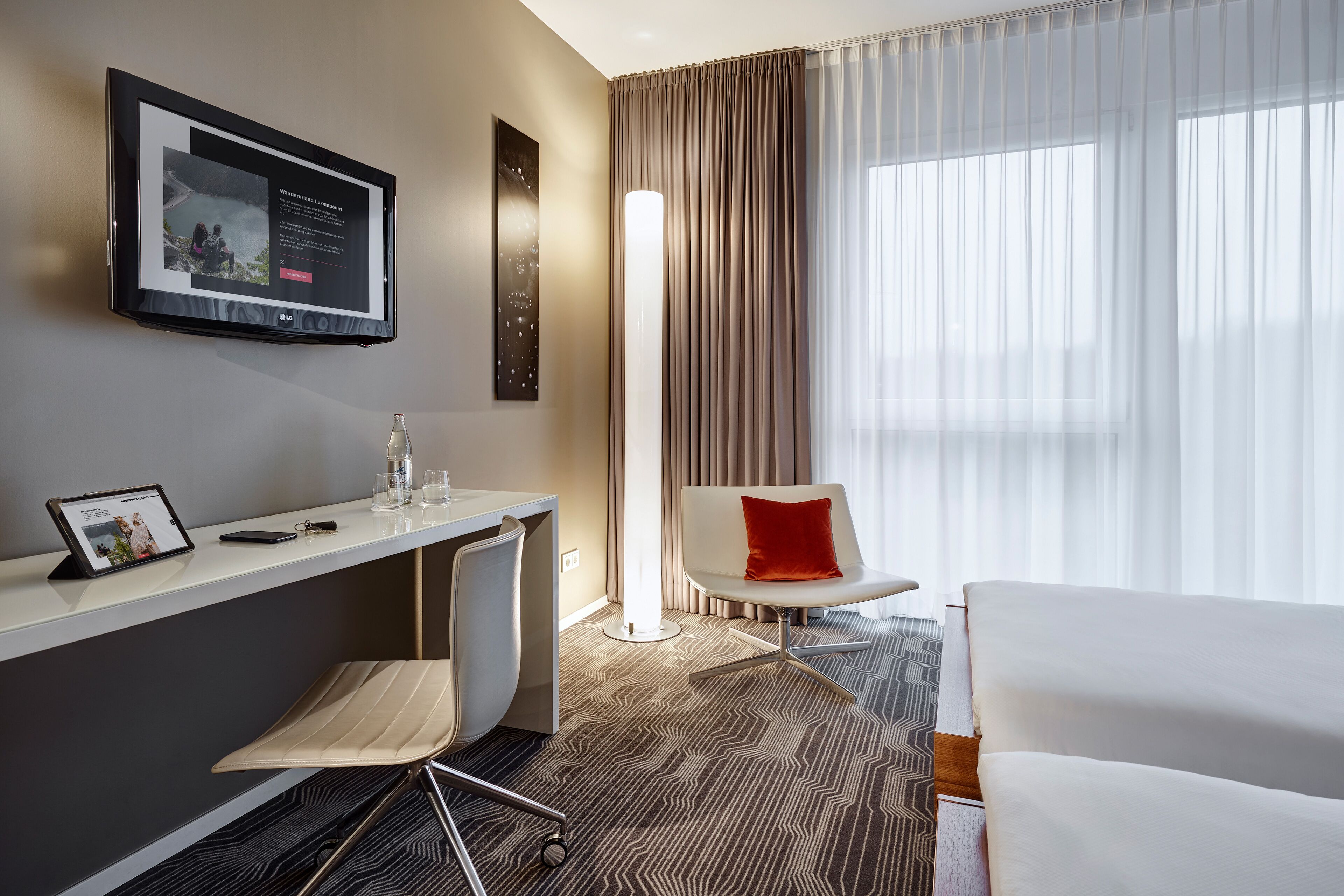 A modern hotel room with a desk, chair, bed, and a large window with curtains.