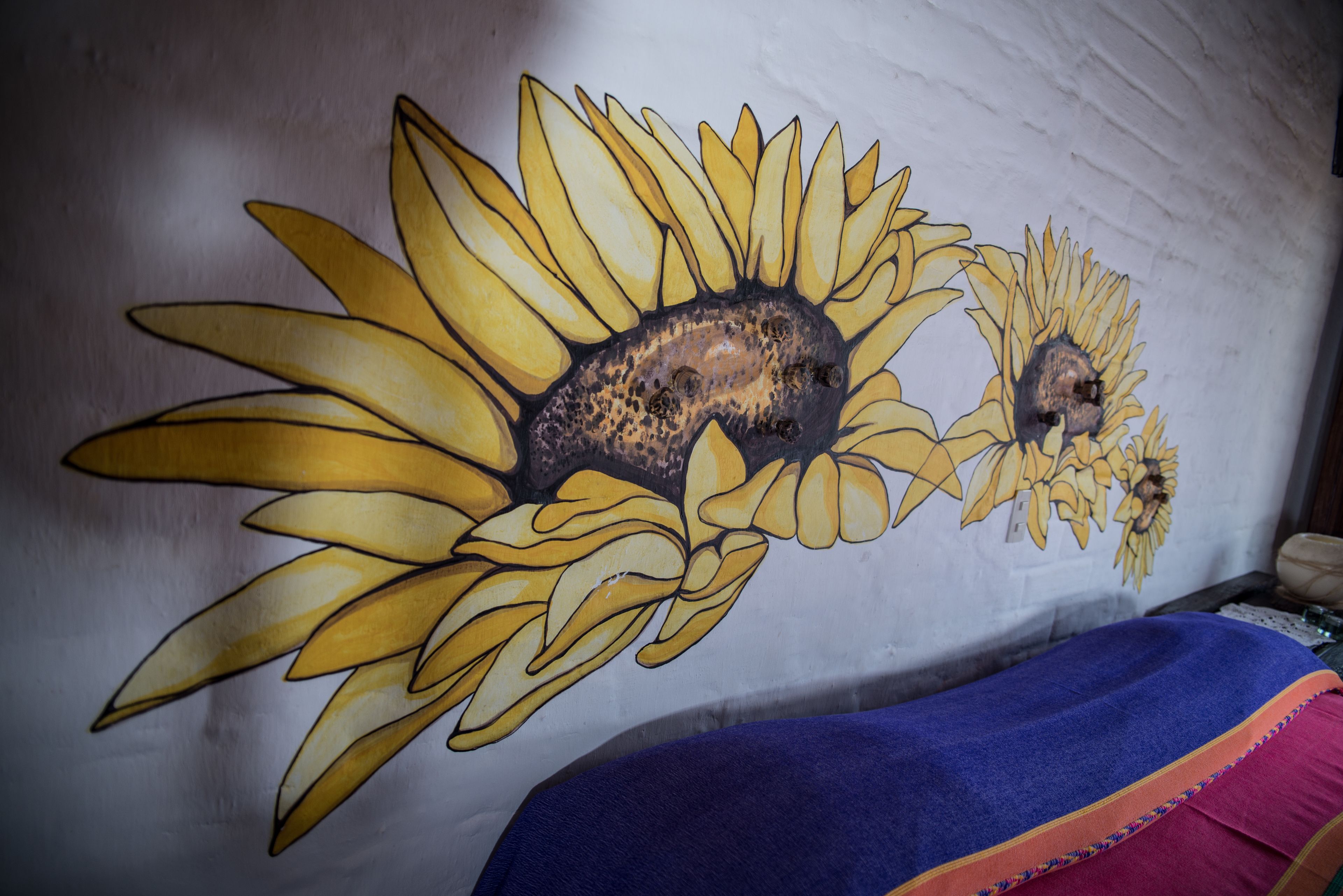 A wall with a sunflower mural, a bed with a blue and pink blanket, and a small table with a lamp.