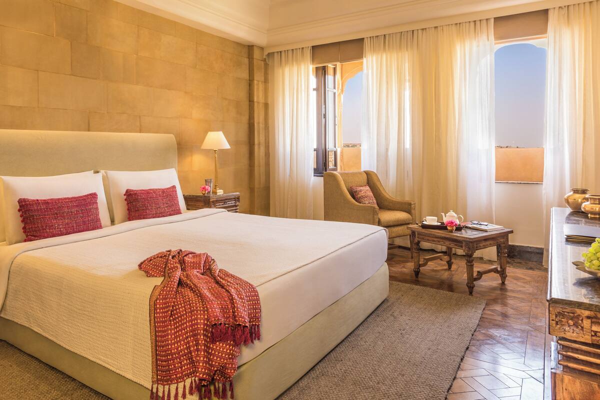 Habitación de hotel con una cama grande, dos mesitas de noche, una silla, una pequeña mesa con un florero y vistas al exterior a través de una ventana.
