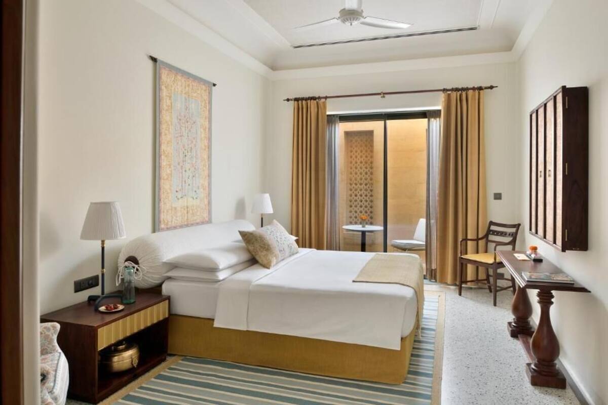 Habitación de hotel con cama, mesitas de noche, lámpara, silla, mesa y vistas por una puerta.