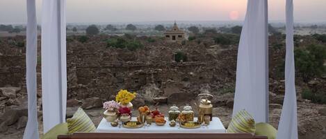 A dining setup with a table and cushions, overlooking ancient ruins and a sunset.