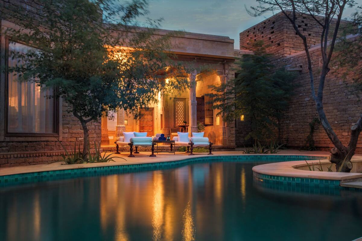 Zona de piscina con dos sillas reclinables y una mesa, rodeada por un muro de piedra y árboles.