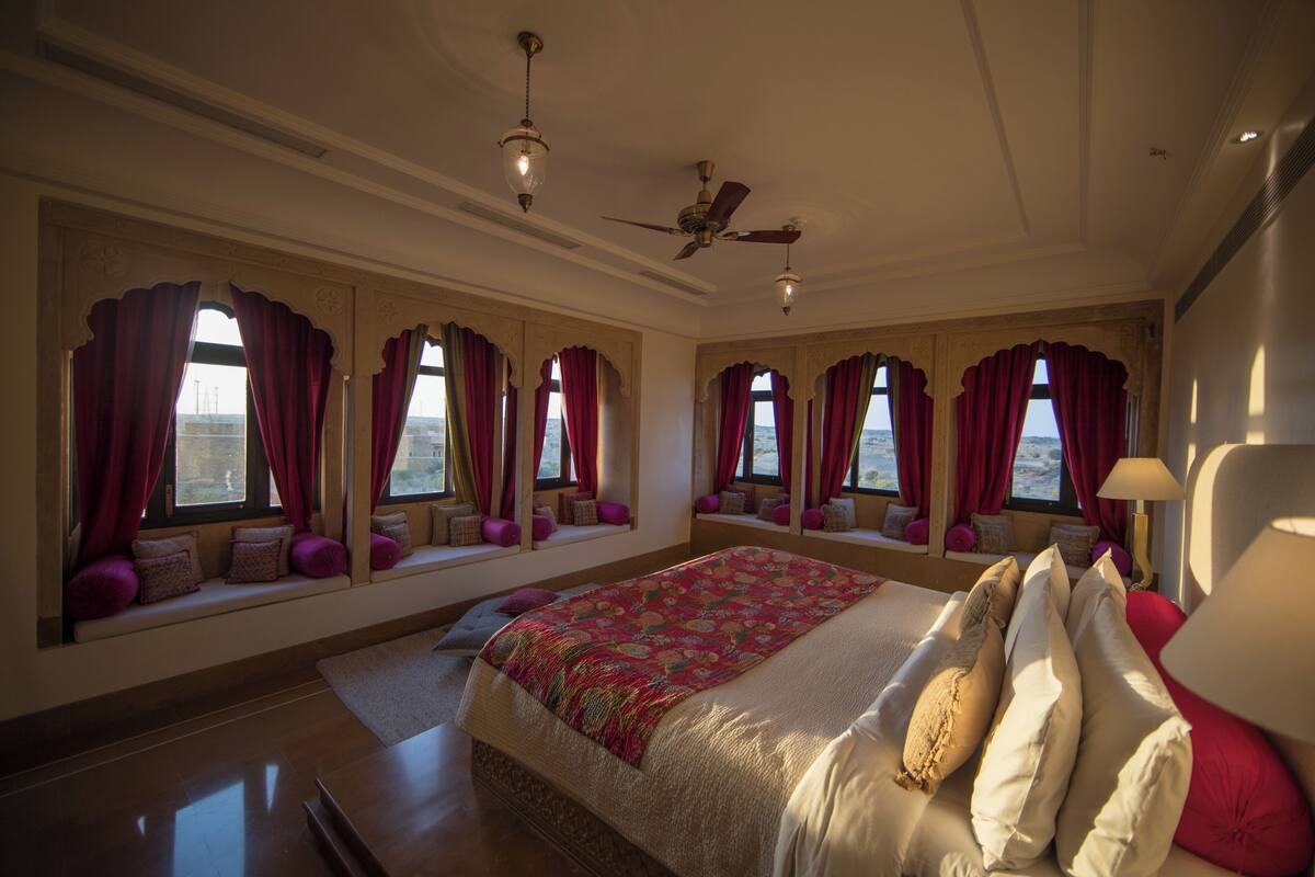 Un dormitorio amplio con una cama grande, varias ventanas con cortinas rojas, un ventilador de techo y vistas al exterior.