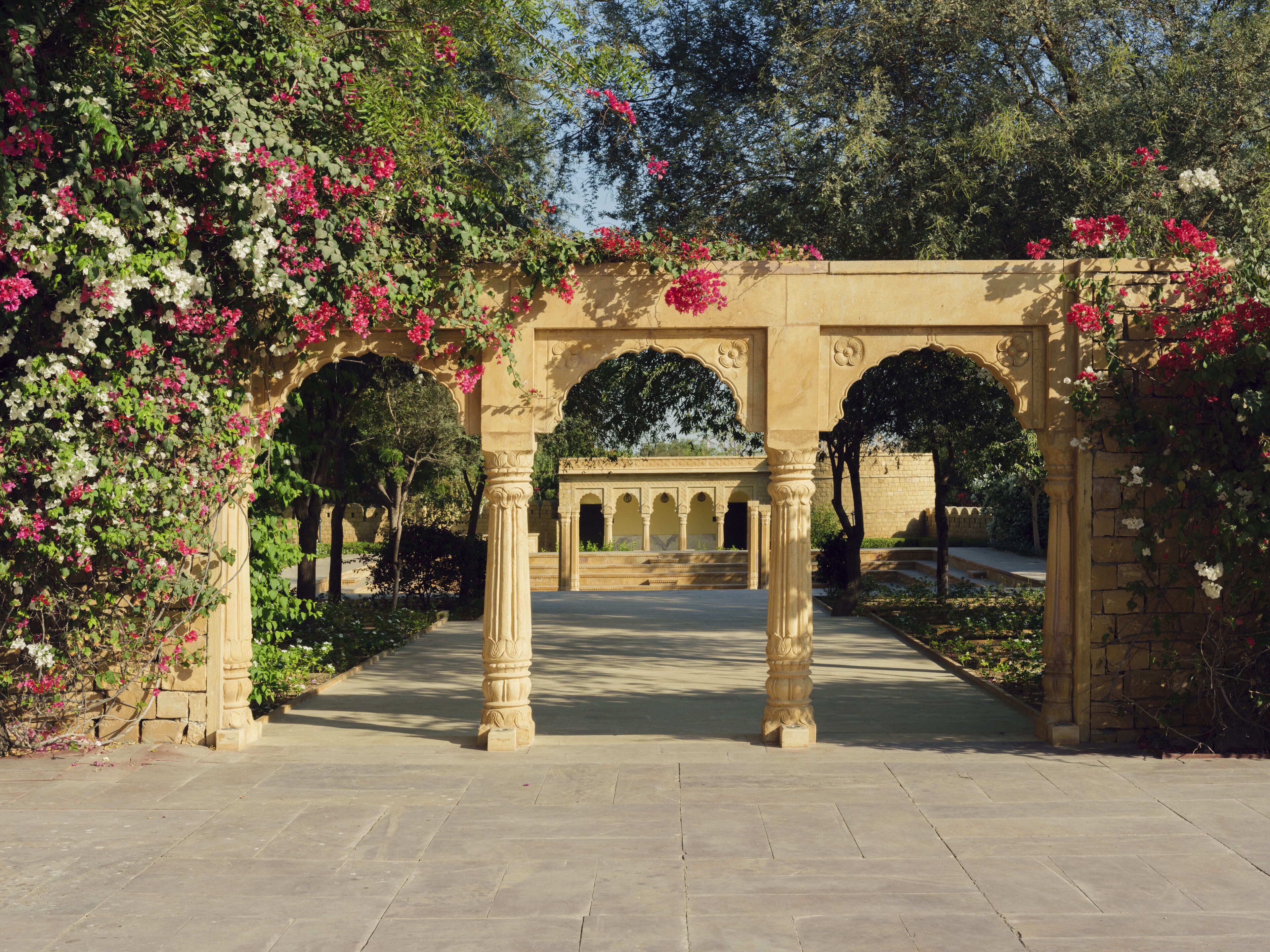 A stone archway with intricate carvings, leading to a courtyard with a tiled floor and lush greenery.