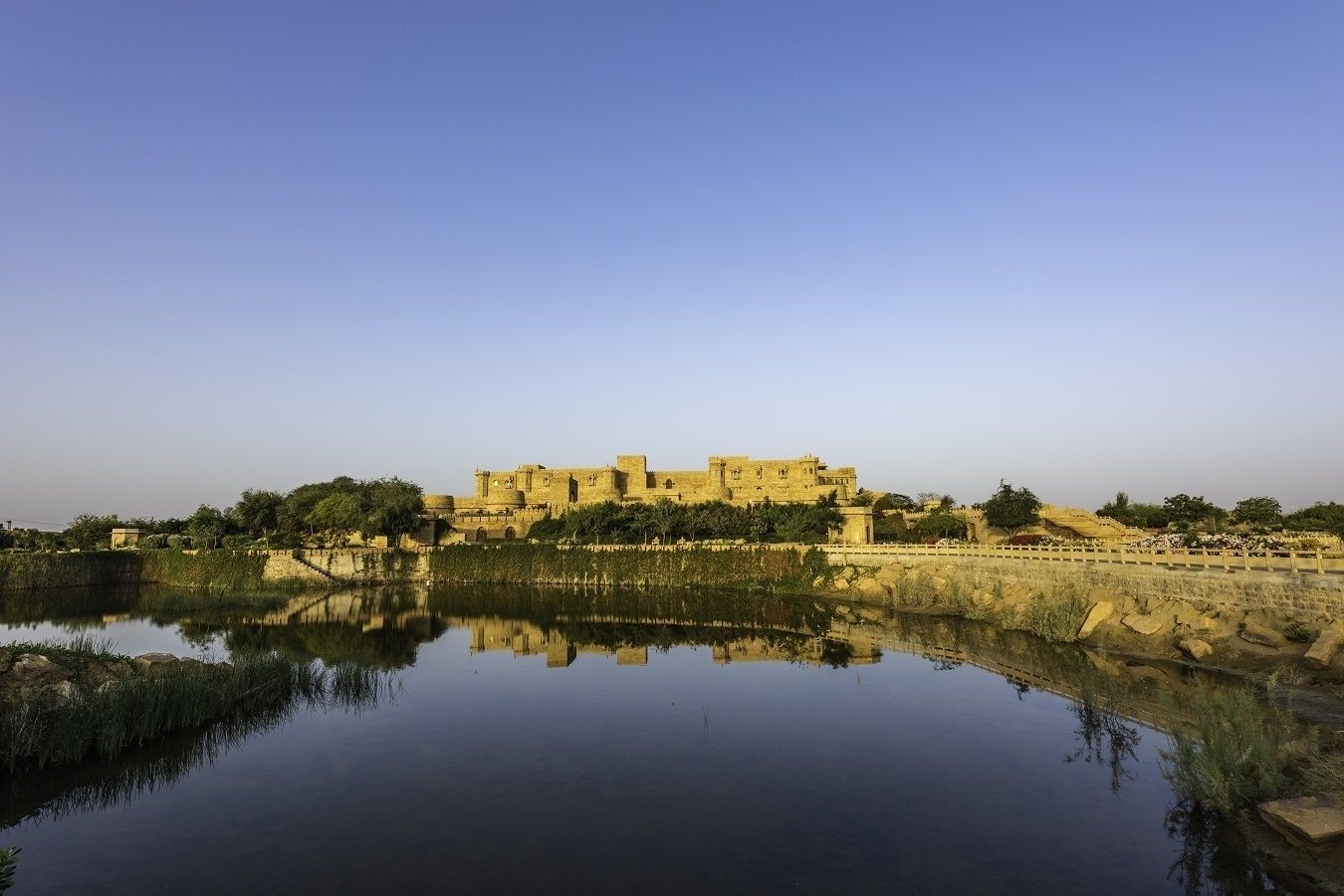 Una fortaleza histórica reflejada en aguas tranquilas, rodeada de vegetación y un cielo despejado.