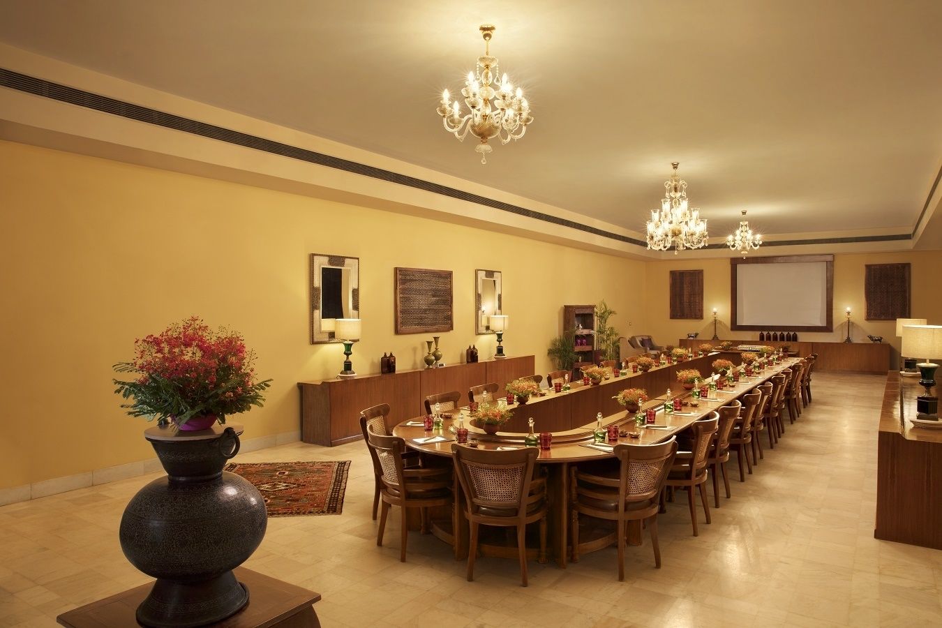 A long dining table with chairs, chandeliers, and a projector screen on the wall.