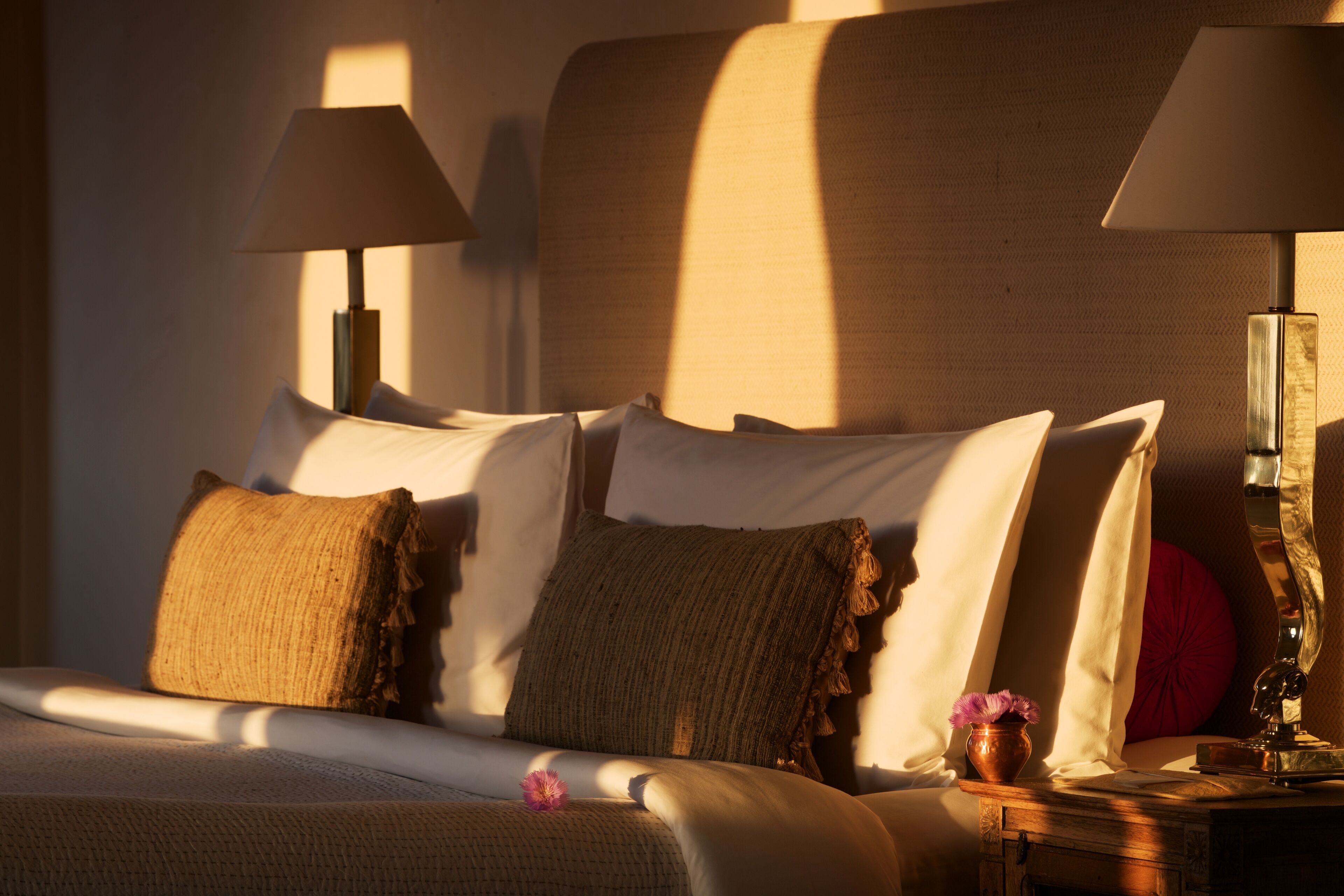 A neatly made bed with pillows and a vase of flowers, two lamps, and a wooden nightstand.