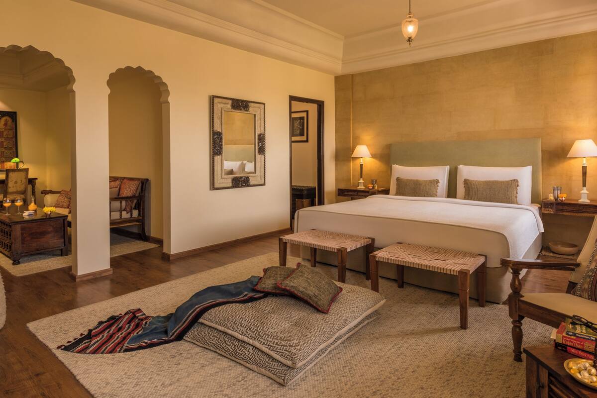 Una habitación de hotel amplia con una cama grande, dos mesitas de noche con lámparas, un banco de madera, una alfombra estampada y un espejo decorativo.