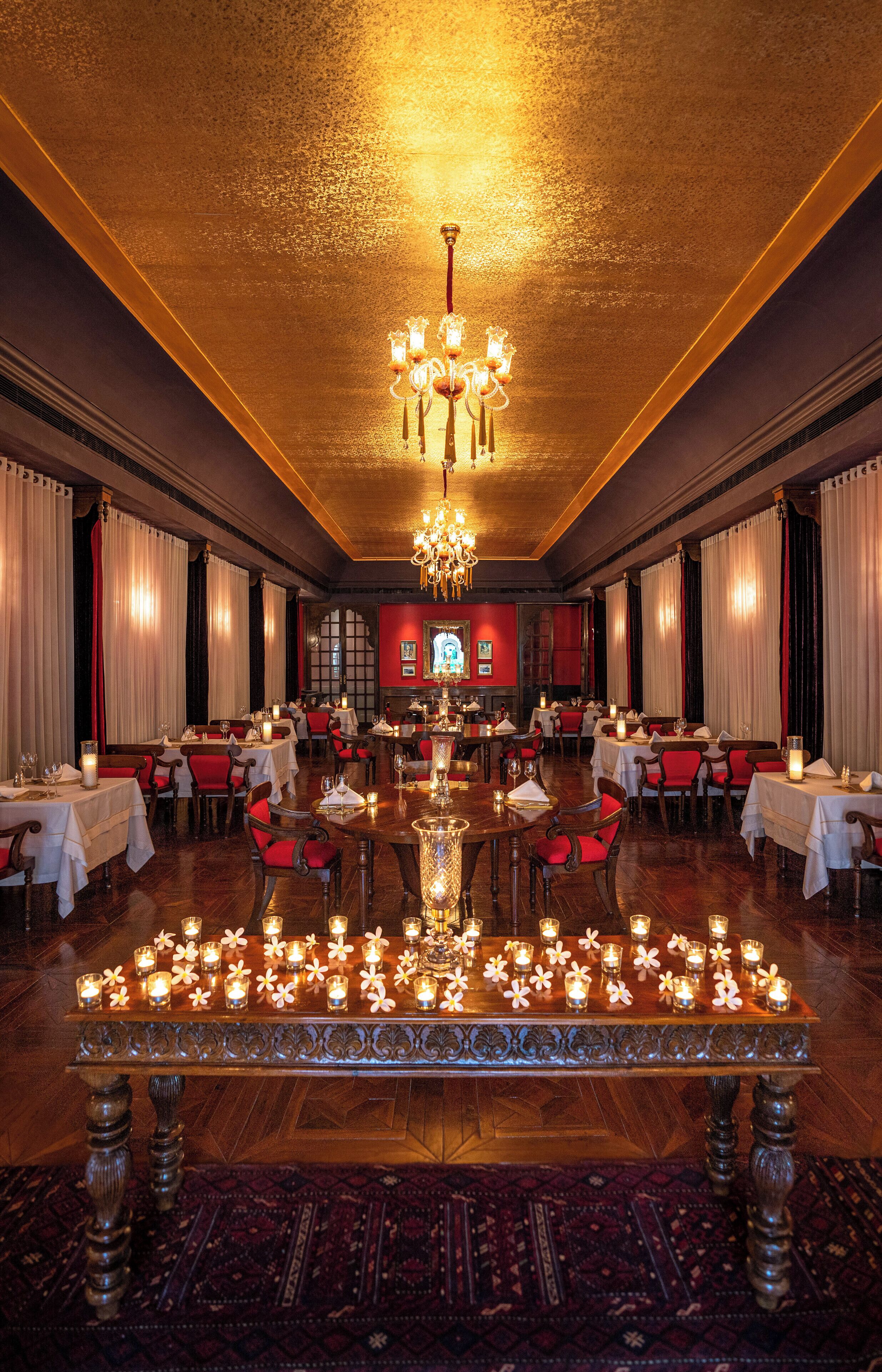 A formal dining room with tables set for multiple guests, a chandelier, and candles on a central table.