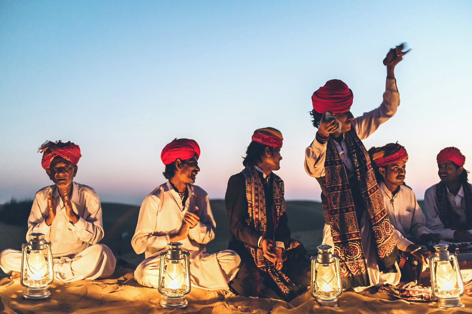 A group of people sitting on the ground with lanterns, dressed in traditional attire, in an outdoor setting.