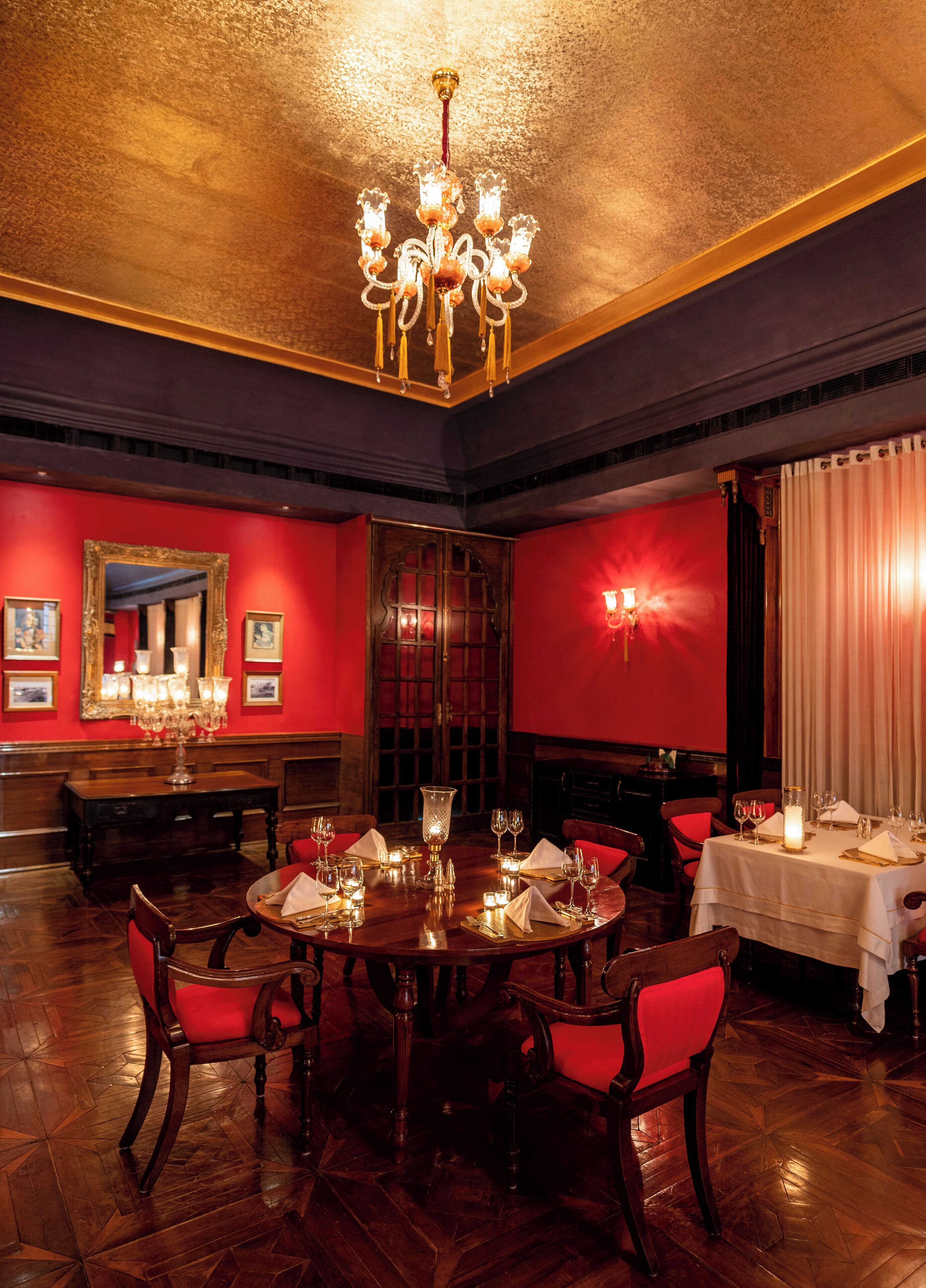 A formal dining room with a round table set for four, red upholstered chairs, a chandelier, and a mirror on the wall.