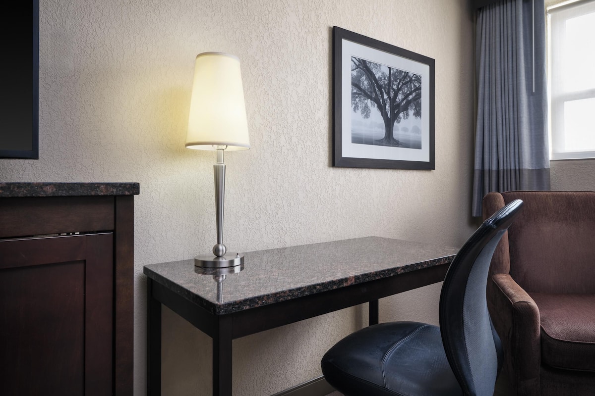 A hotel room with a desk, a chair, a lamp, a framed picture on the wall, and a window with curtains.