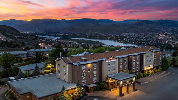 Un hotel de varios pisos, con estacionamiento y vistas a un río y montañas al fondo.