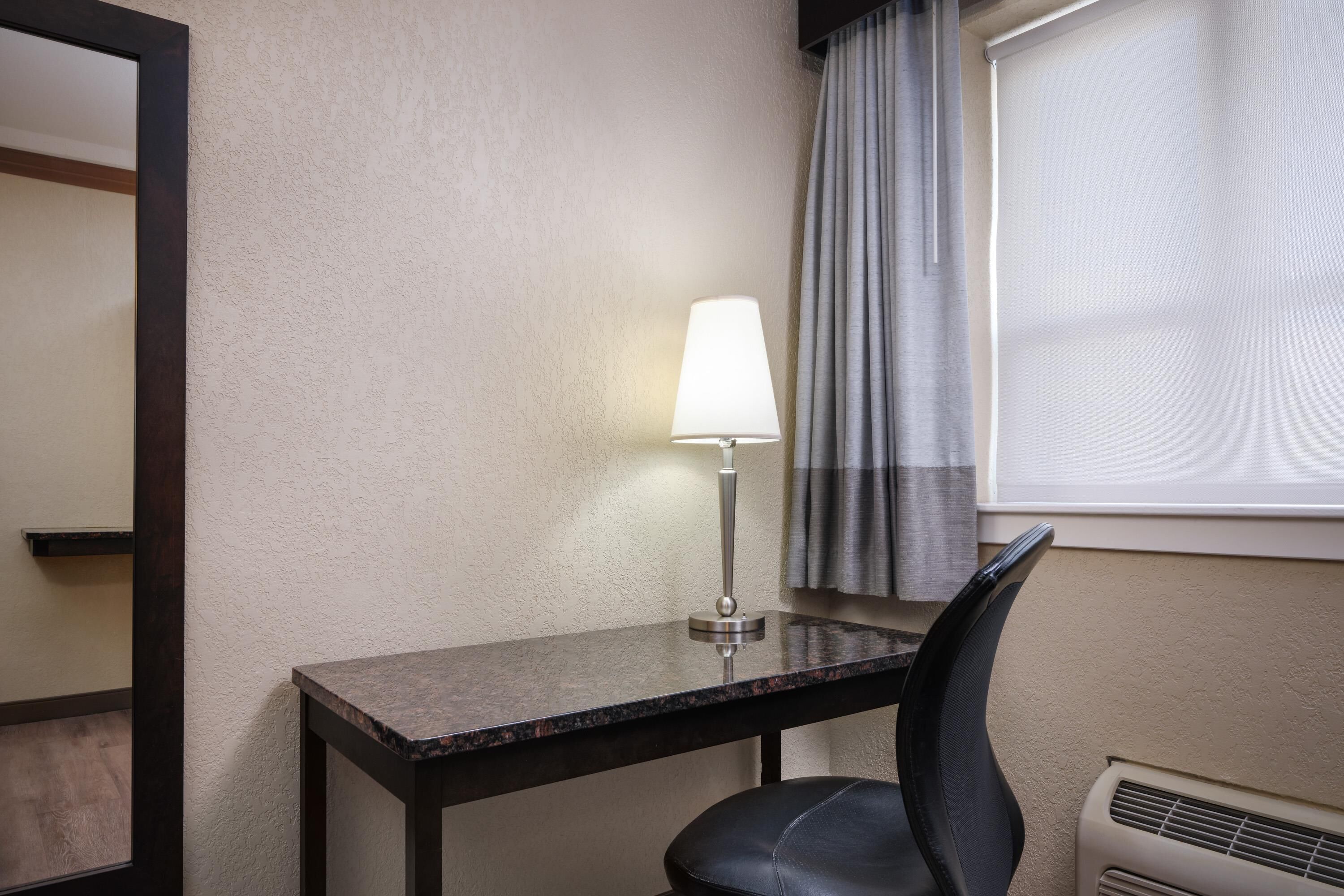 A hotel room with a desk, a chair, a lamp, a window with curtains, and a wall-mounted air conditioner.