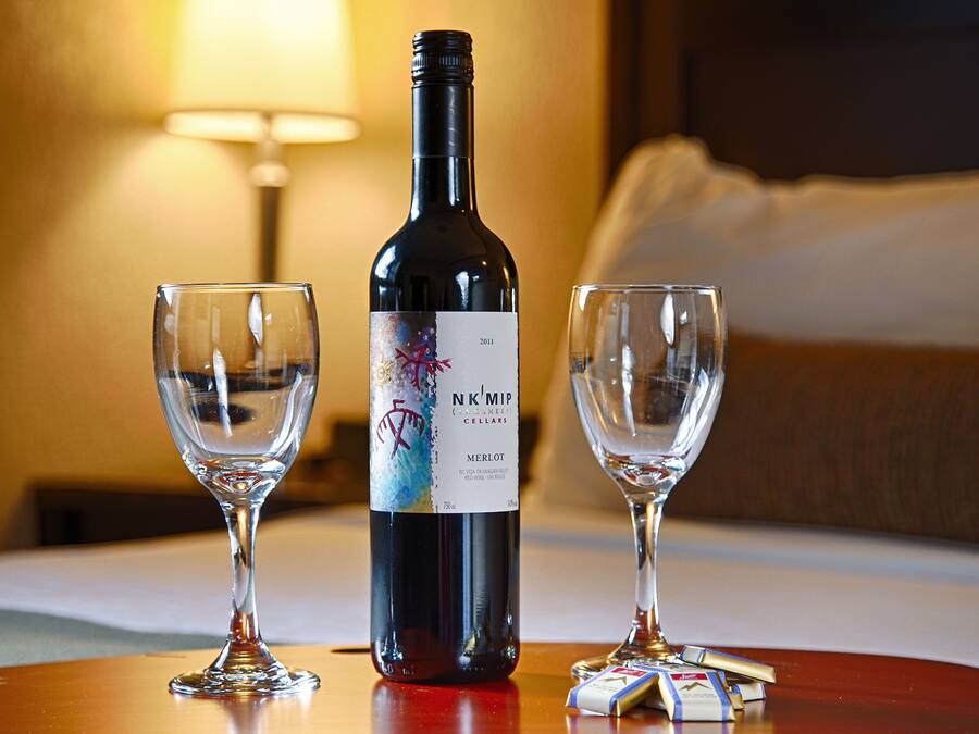 A bottle of Merlot wine, two empty wine glasses, and a small packet of cigarettes on a wooden table in a hotel room.