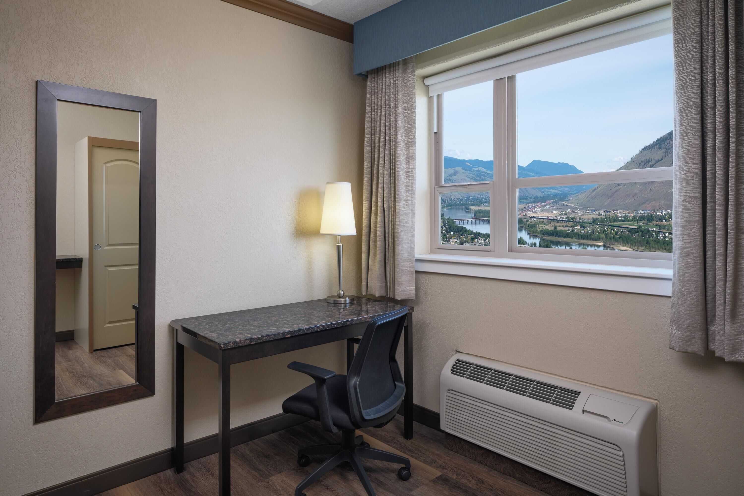 Une chambre d’hôtel équipée d’un bureau, d’une chaise, d’une lampe, d’une fenêtre avec des rideaux, et offrant une vue sur un lac et des montagnes.
