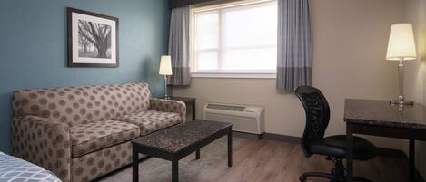 A hotel room with a patterned sofa, a small table, a desk with a chair, a window with curtains, and a framed picture on the wall.