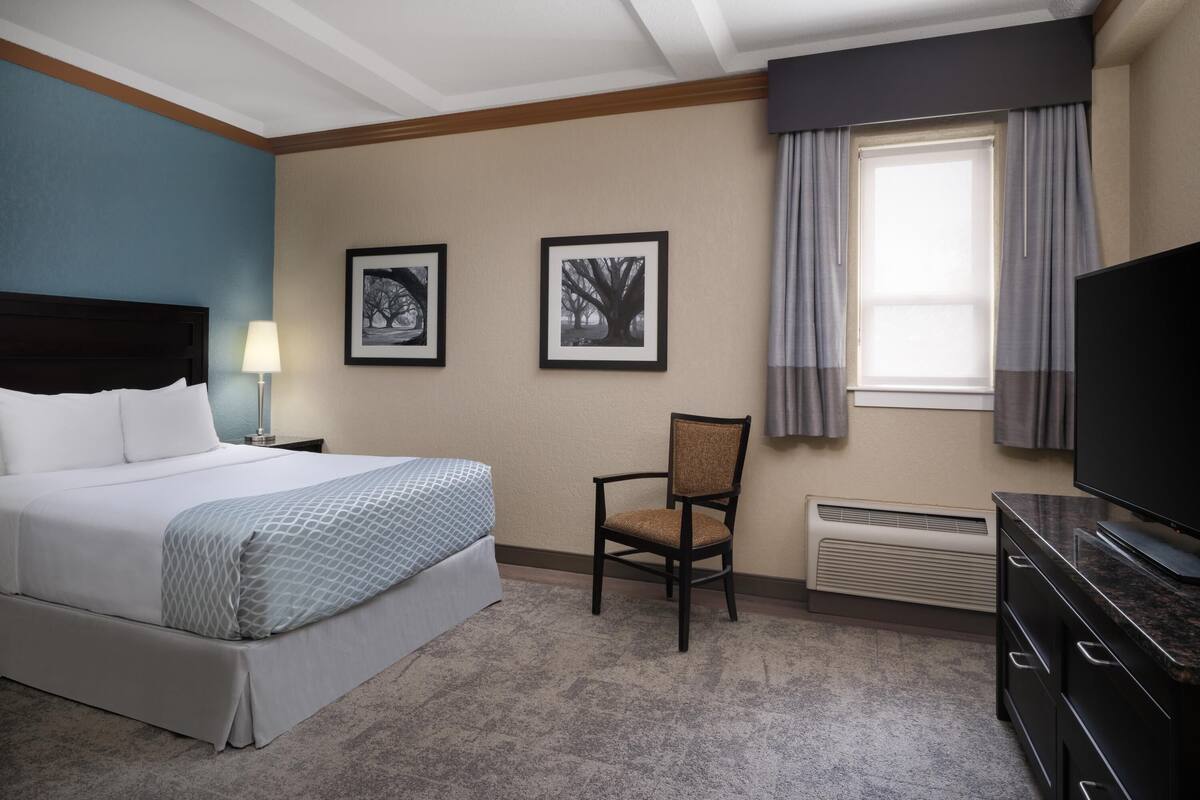 A hotel room with a bed, a chair, a TV, and framed pictures on the wall.