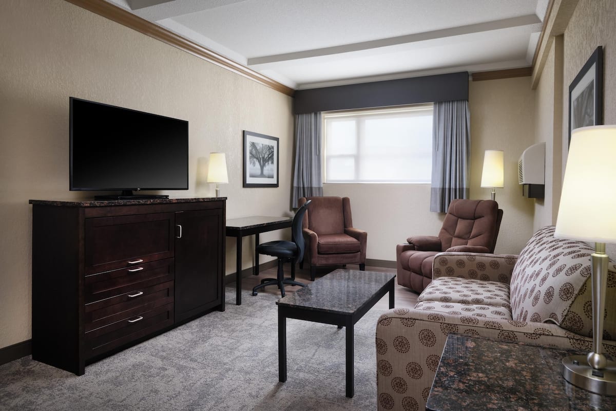 A hotel room with a flat-screen TV, a dark wooden cabinet, a patterned sofa, a small coffee table, and a desk with a chair.