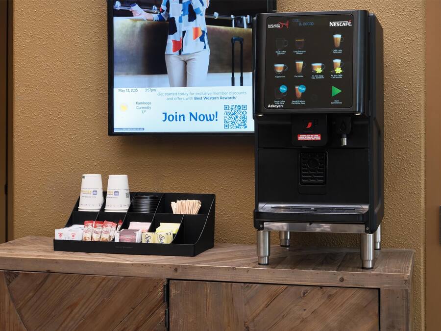 A coffee machine with a touchscreen interface, a tray of coffee cups, and a promotional screen on the wall.
